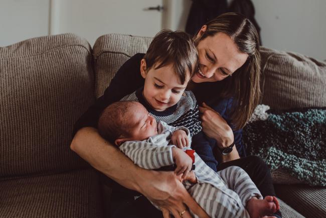 Mother sitting on the couch with two boys on her lap with Lifestyle Newborn Photographer Perth