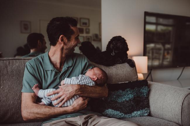 Father holding new baby with dog on the back of the couch with Lifestyle Newborn Photographer Perth