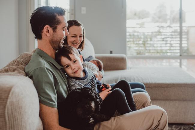 Little boy sitting on his dad's lap and smiling at the camera with Lifestyle Newborn Photographer Perth