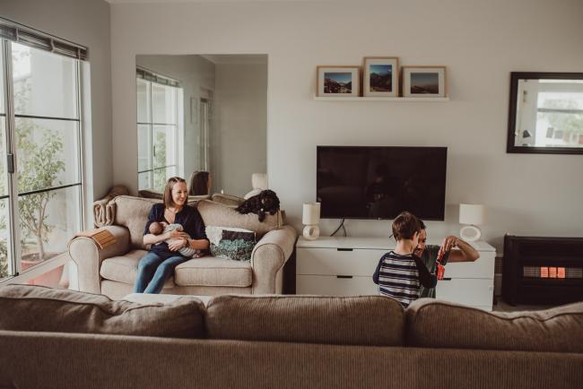 Mother feeding new baby on the couch as father and son play on the floor with Lifestyle Newborn Photographer Perth