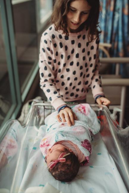 Older sister reaching into the bassinet of her new baby sister with Perth Fresh 48 Photographer at SJOG Mt Lawley