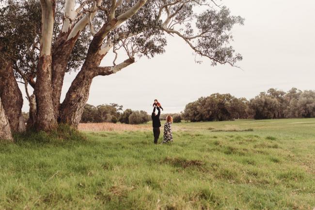 Father lifting his daughter into the sky as mother stands next to them under a tree with Perth family photographer at Perry's Paddock
