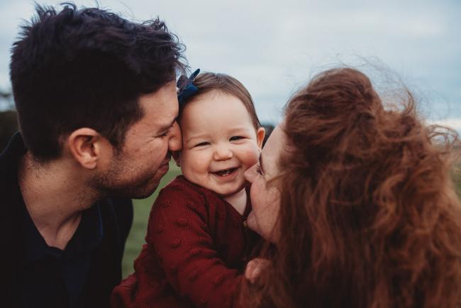 Parents kiss baby daughter who is smiling with Perth family photographer at Perry's Paddock