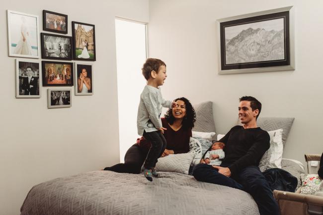 Little boy jumping on the bed in front of his parents and baby brother with lifestyle newborn photographer Perth