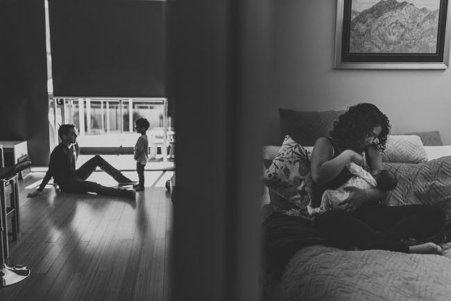 Black and white image of father and son on the floor whilst mum breastfeeds baby with lifestyle newborn photographer Perth