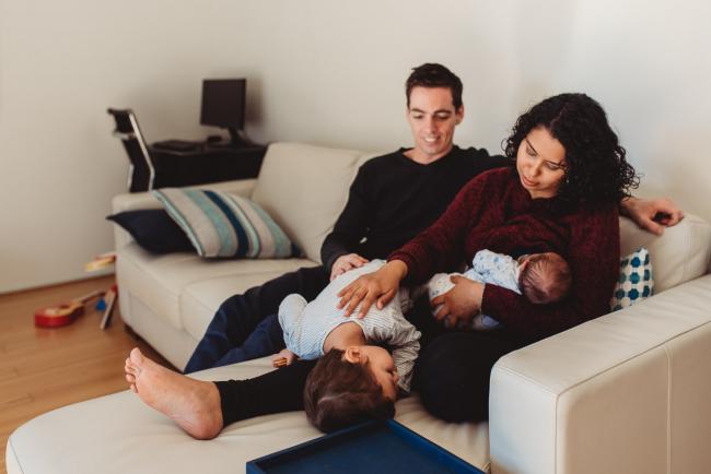 Little boy laying across his mother's legs as she feeds his baby brother with lifestyle newborn photographer Perth