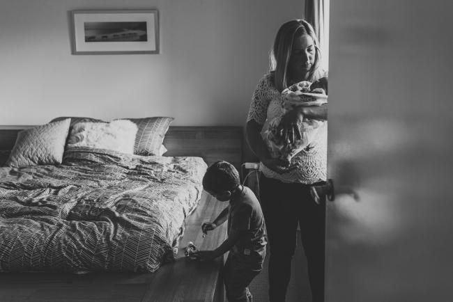 Black and white image of mother standing and holding her new baby while her son plays with his trucks next to her with Perth newborn lifestyle photographer