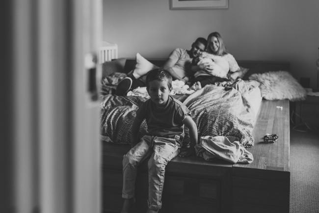 Perth-Newborn-Photographer-45-of-60 black and white image of little boy sitting on the edge of the bed with parents and newborn behind him with Perth newborn lifestyle photographer