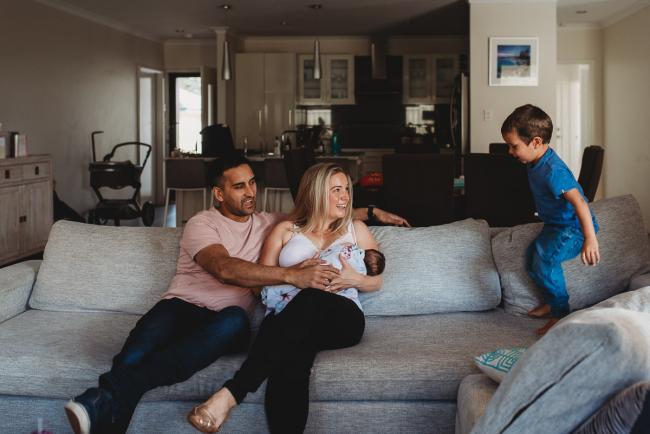 Perth-Newborn-Photographer-5-of-60 Boy jumping on the couch next to his parents who are holding his baby sister with Perth newborn lifestyle photographer