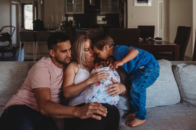 Little boy kissing his baby sister who is being held by his parents on the couch with Perth newborn lifestyle photographer