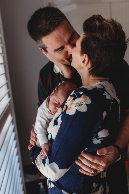Parents kissing as mother holds sleeping baby with Perth newborn lifestyle photographer