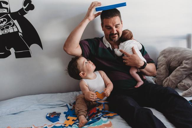 Father holding newborn baby and putting a book on his head as toddler looks up at him with Perth newborn lifestyle photographer