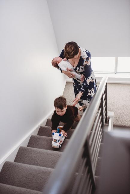Mother carrying newborn upstairs with toddler son pushing his truck with Perth newborn lifestyle photographer