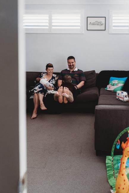 Mother feeding newborn baby on couch while father lifts toddler on his legs next to her with Perth newborn lifestyle photographer