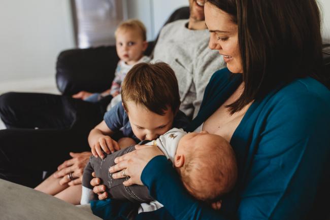 Mother holding new baby as older son reaches over to kiss him with lifestyle newborn photographer Perth