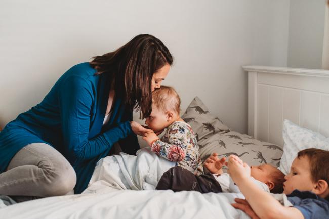 Mother kissing daughter as her three kids are in bed with lifestyle newborn photographer Perth