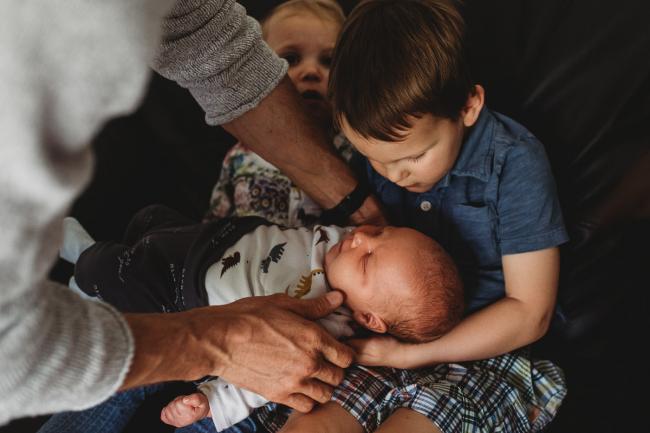 Father placing new baby in older son's arms with lifestyle newborn photographer Perth