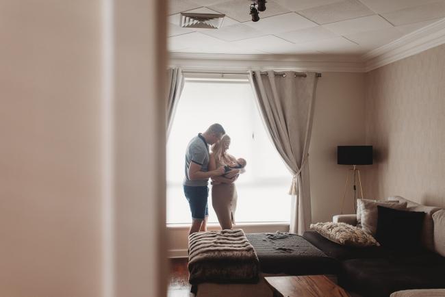 Parents standing in front of window with newborn baby with in-home lifestyle newborn photographer Perth