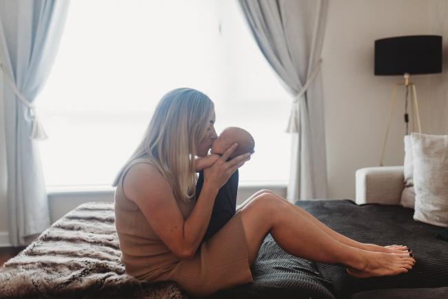 Mother kissing newborn baby as she sits on couch in front of window with lifestyle newborn photographer Perth