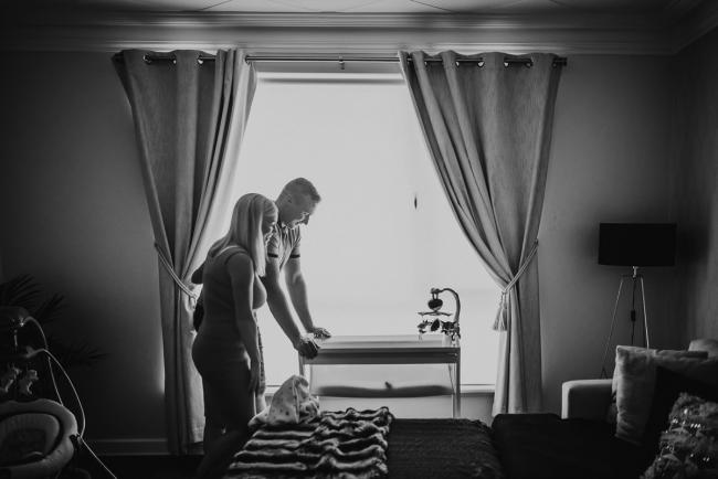 black and white image of mother and father in front of bassinet by window with lifestyle newborn photographer Perth