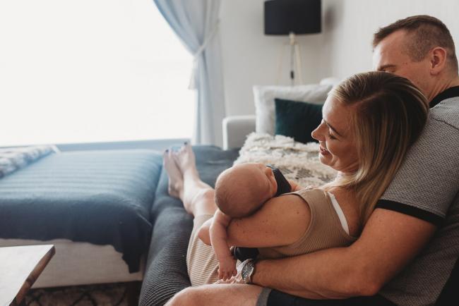 Lifestyle-Newborn-Photographer-Perth-9-of-69 Mother leaning against husband as she smiles down at her newborn baby in her arms with in-home lifestyle newborn photographer Perth