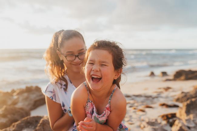 Family-Photographer-Perth-Burns-Beach-19-of-57 Little sister laughing on big sister's lap at Burns Beach with Perth Family Photographer