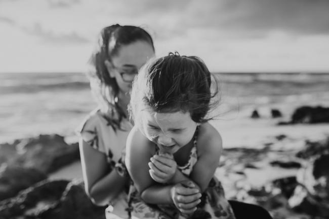 Family-Photographer-Perth-Burns-Beach-20-of-57 black and white image of little girl laughing on sister's lap at Burns Beach with Perth Family Photographer