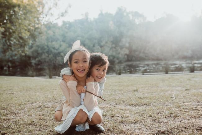 Family-Photographer-Perth-Hyde-Park-17-of-54 Sister sitting on grass as brother hugs her from behind during Perth extended family session at Hyde Park with Perth family photographer