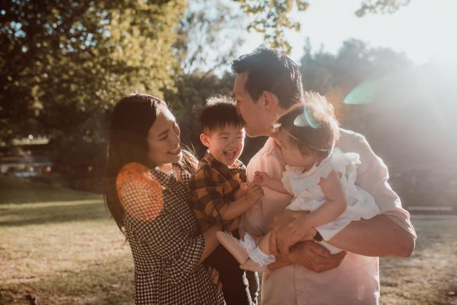 Family-Photographer-Perth-Hyde-Park-4-of-54 Father holding daughter as kissing son who is being held by mother with sunflare during Perth extended family session at Hyde Park with Perth family photographer