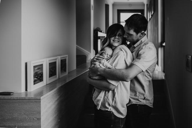 Newborn-Lifestyle-Photographer-Perth-25-of-50 black and white image of mother holding newborn baby with father hugging from behind during Perth lifestyle newborn photography session