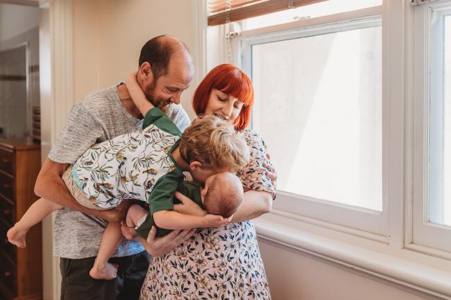 Father holding son who is leaning over to kiss baby brother being held by mother during in-home Perth newborn lifestyle photography session