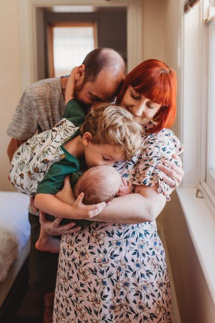 Little boy kissing baby brother who is being held by parents during in-home Perth newborn lifestyle photography session