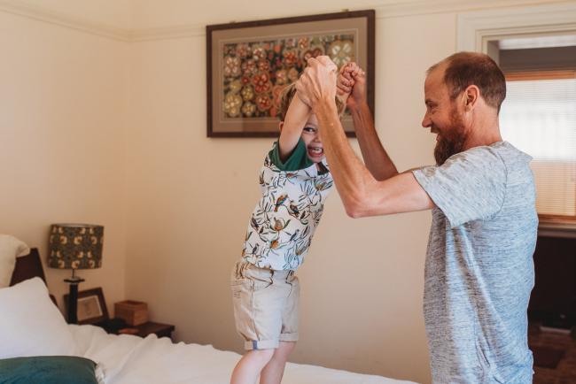Father holding hands with son who is jumping on the bed during in-home Perth newborn lifestyle photography session