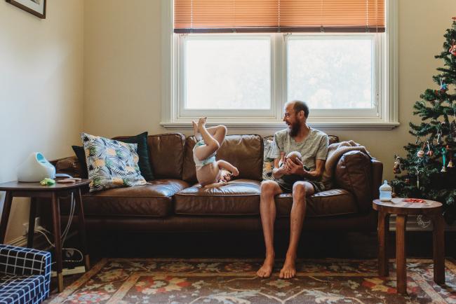 Father holding newborn baby on couch as toddler jumps on couch next to him during in-home Perth newborn lifestyle photography session