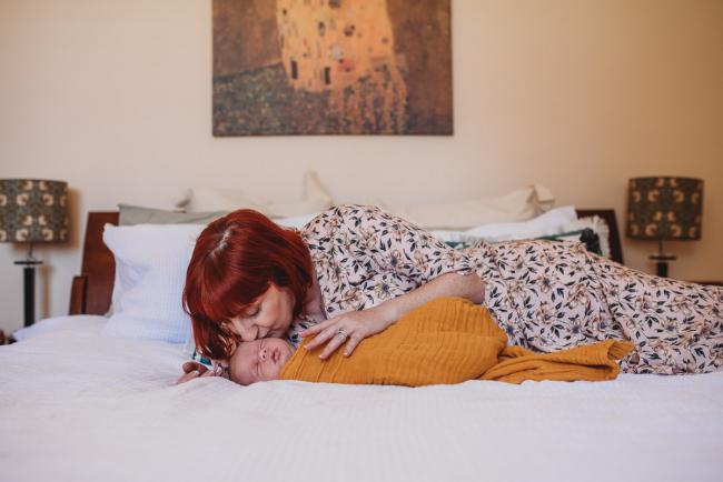Mother laying on bed kissing newborn baby during in-home Perth newborn lifestyle photography session