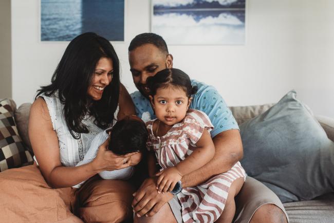 Mother smiling and holding newborn as little girl sits on fathers lap and looks at camera with in-home Perth lifestyle newborn photographer