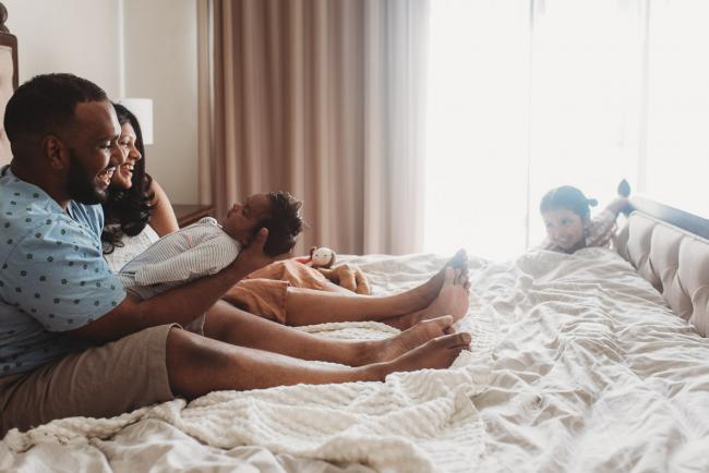Father holding newborn baby on bed as he and mother smile at older daughter with in-home Perth lifestyle newborn photographer