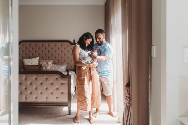 Parents holding newborn baby by window with in-home Perth lifestyle newborn photographer