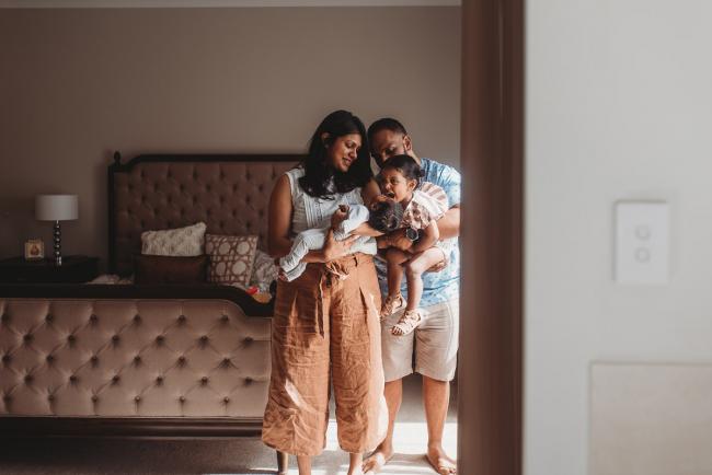 Parents holding daughter and newborn baby by window with in-home Perth lifestyle newborn photographer