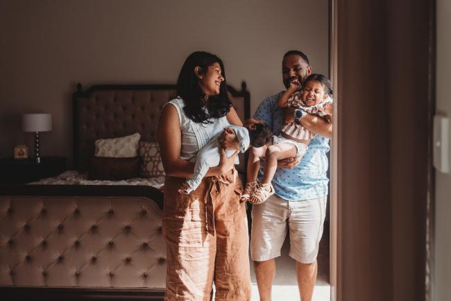 Parents holding daughter and newborn baby by window with in-home Perth lifestyle newborn photographer