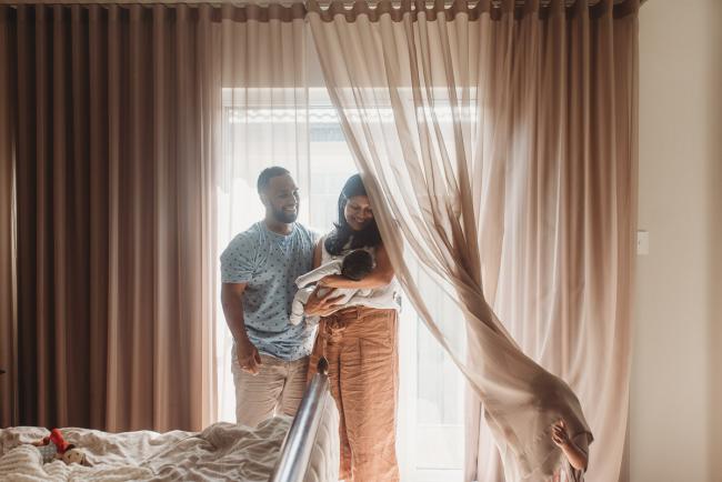 Parents holding newborn baby by window as daughter plays in curtains with Perth newborn photographer