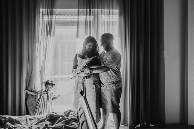 black and white image of parents holding newborn baby by a window as daughter plays in the curtains with in-home Perth lifestyle newborn photographer