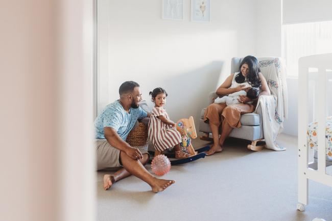 Mother breastfeeding newborn baby as father and daughter play on the floor nearby with in-home Perth lifestyle newborn photographer