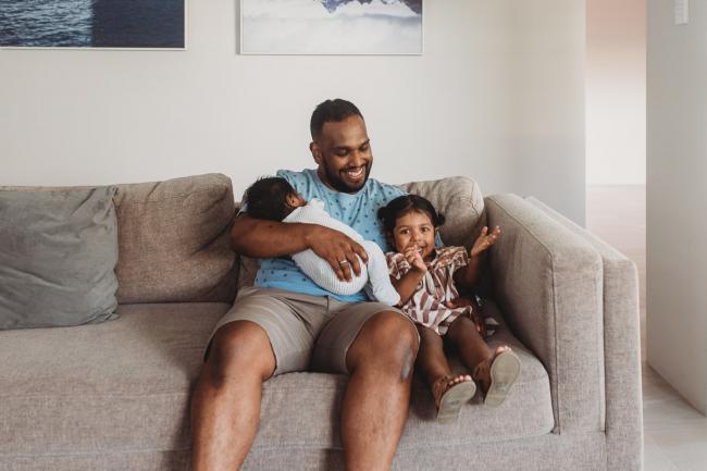 Father holding newborn baby and daughter on couch with in-home Perth lifestyle newborn photographer