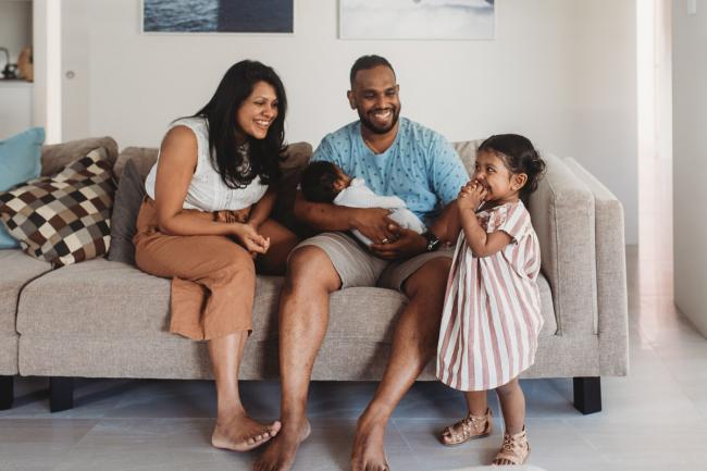 Parents holding newborn baby on couch as daughter stands next to them with in-home Perth lifestyle newborn photographer