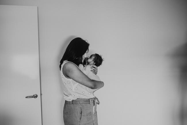 Black and white image of mother standing against the wall with newborn baby with in-home Perth lifestyle newborn photographer