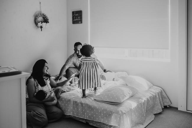 Black and white image of little girl jumping on bed towards father, as mother and newborn baby sit on the floor with in-home Perth lifestyle newborn photographer