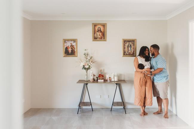 Parents standing next to religious wall holding newborn baby with in-home Perth lifestyle newborn photographer