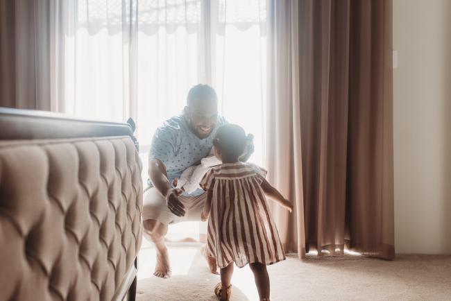 Father holding newborn baby as squatting and holding his arms out for daughter to run into them with in-home Perth lifestyle newborn photographer