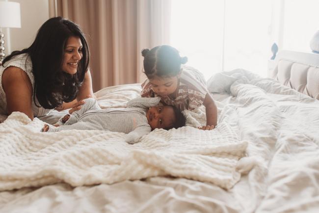 Mother leaning on bed as she watches her daughter kiss her newborn baby with in-home Perth lifestyle newborn photographer